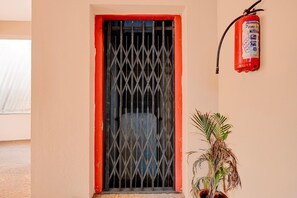 Una ventana con una reja metálica y un marco rojo, un extintor colgado en la pared, y una planta en maceta junto a la ventana.