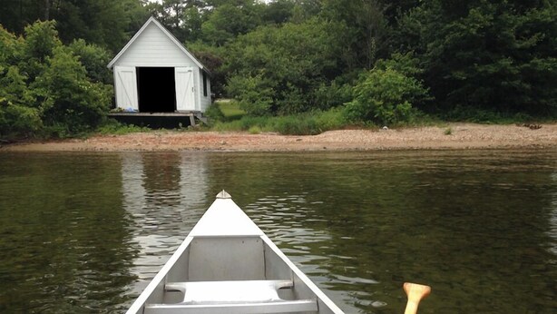 Een kano op kalm water met een houten peddel, een wit gebouw aan de oever en weelderige begroeiing op de achtergrond.