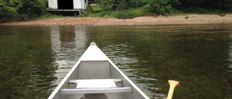 A canoe on calm water with a wooden paddle, a white building on the shore, and dense greenery in the background.