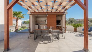 En udendørs terrasse med et træpergola, et spisebord med stole, og et køkkenområde kan anes gennem konstruktionen.