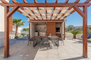 An outdoor patio with a wooden pergola, a dining table with chairs, and a kitchen area visible through the structure. - Modern Escape Villa with Private Pool (Solin)