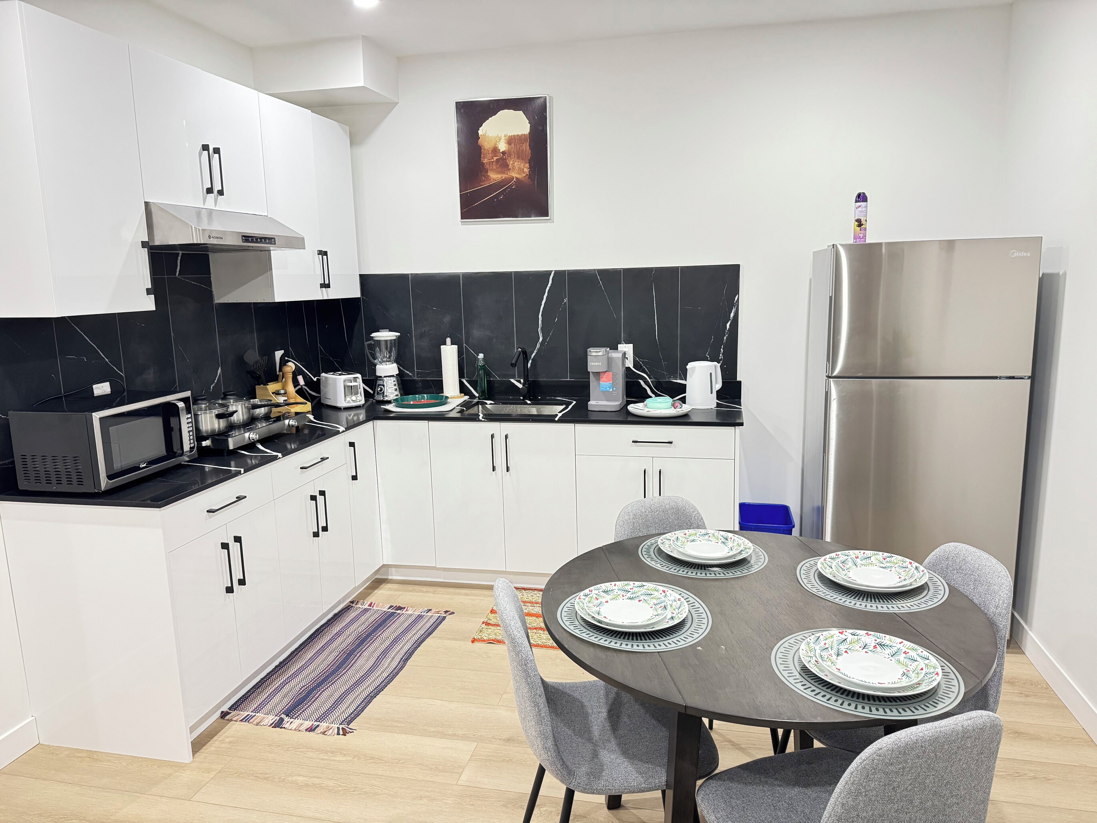 A modern kitchen with white cabinets, a black countertop, a stainless steel refrigerator, and a dining area with a round table and grey chairs.