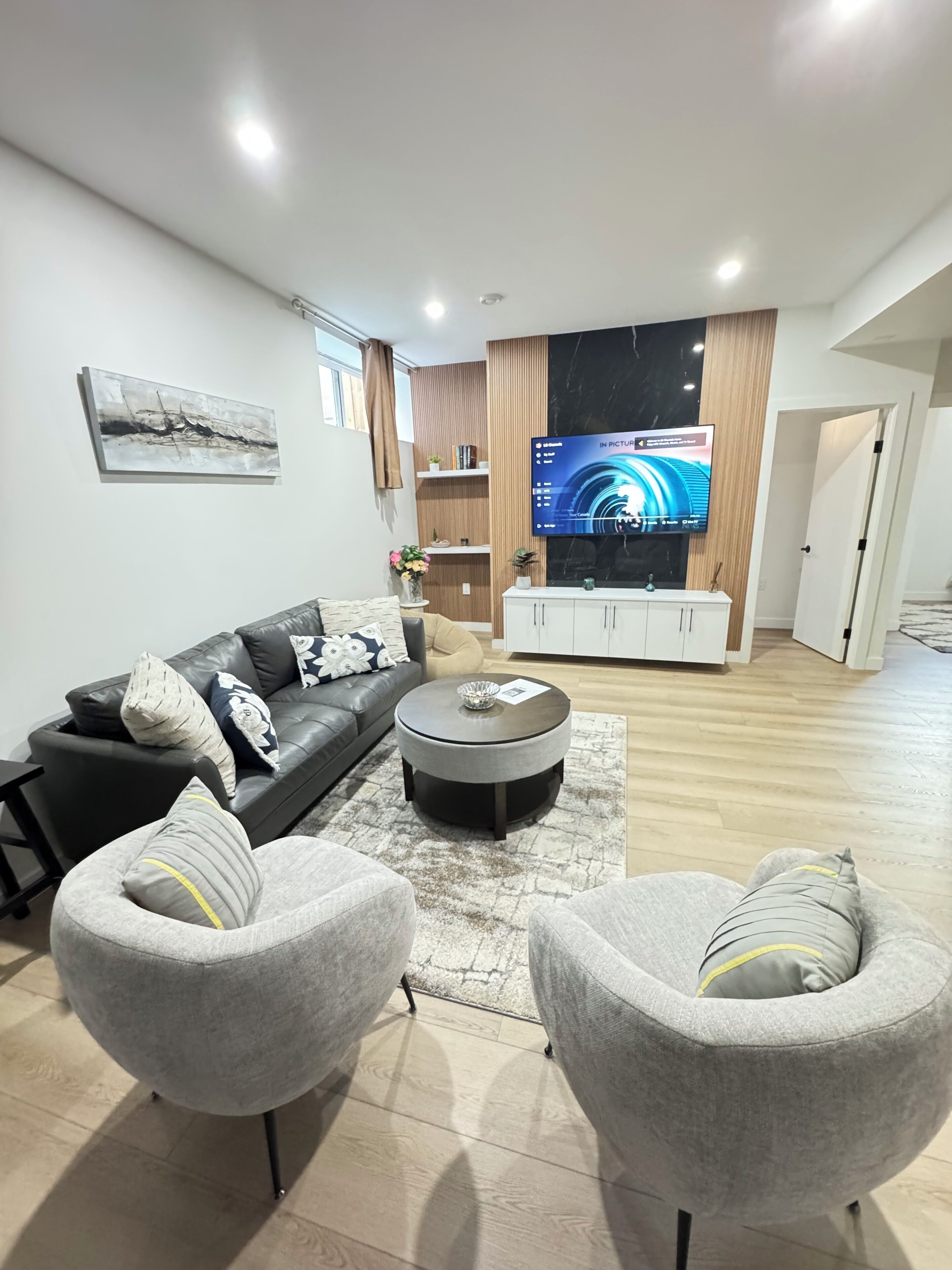 Ein modernes Wohnzimmer mit einem Ledersofa, grauen Sesseln, einem Couchtisch und einem flachbildfernseher, der an einer Holzwand befestigt ist.