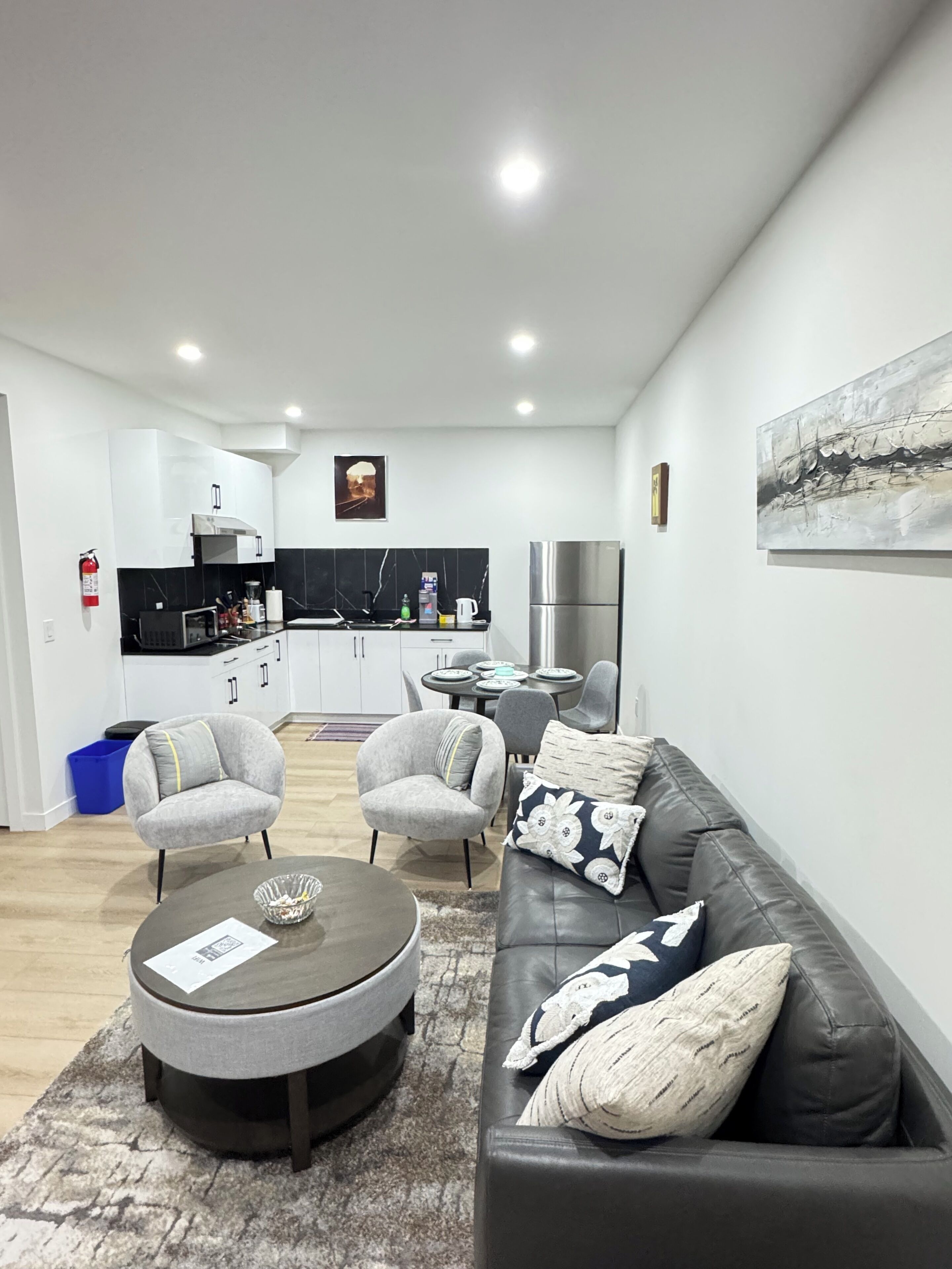 A modern living room with a black leather sofa, grey armchairs, a coffee table, and a dining area with a small table and chairs.