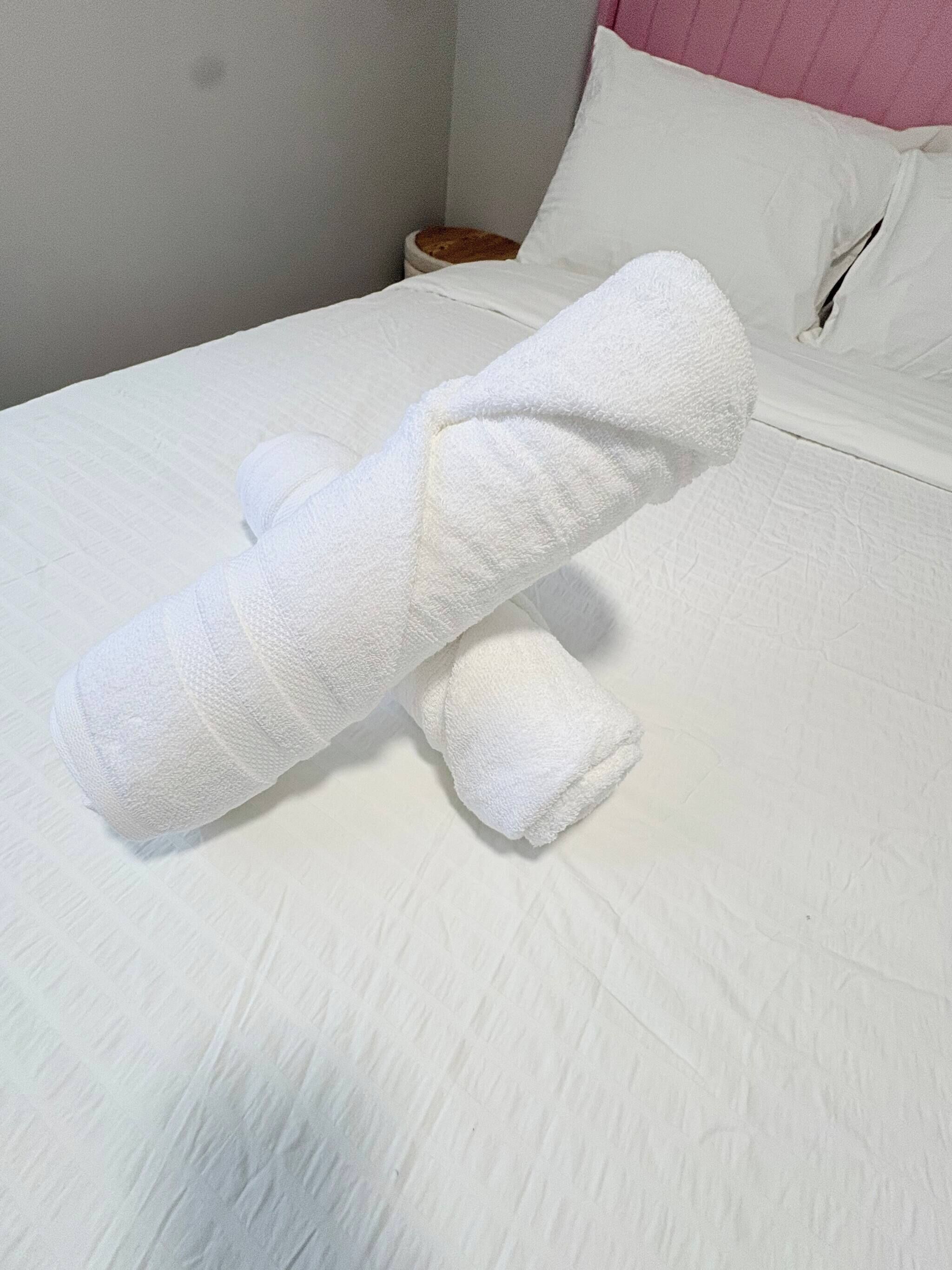 A bed with white bedding, a pillow, and a rolled-up towel.