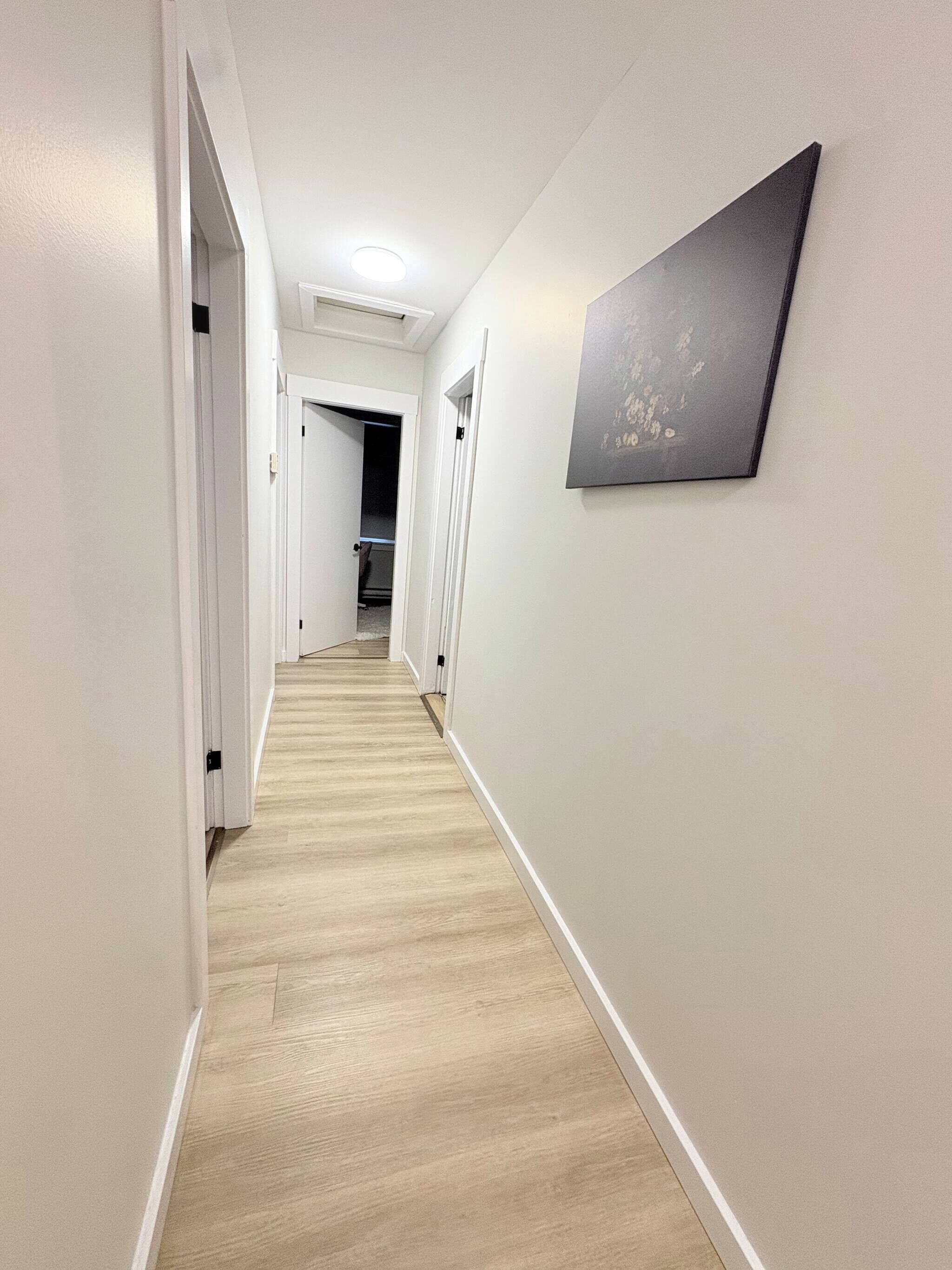 A hallway with a light wooden floor, white walls, and a framed picture on the right.