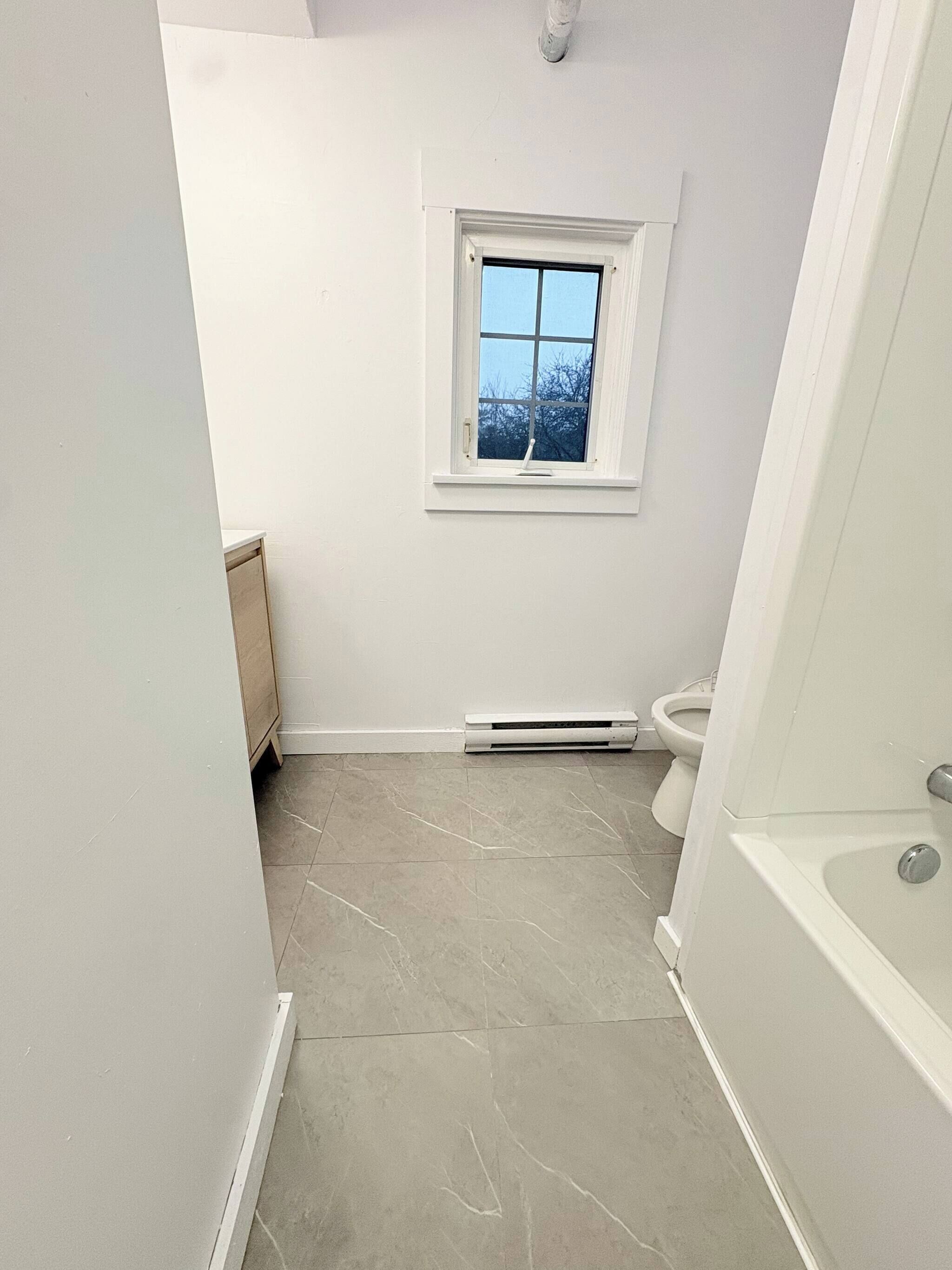 A bathroom with a window, a toilet, a bathtub, and a heater on the floor.