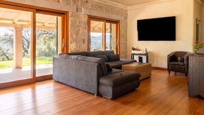 Amplia sala de estar con pared de piedra, un sofá grande, televisor y vistas al exterior.