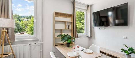 A modern room with a dining table, chairs, a TV, and a window with a view of greenery.