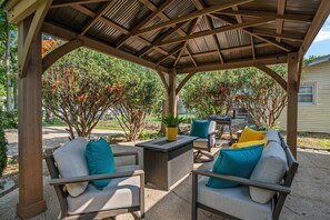 A covered outdoor seating area with wicker furniture and cushions, surrounded by trees and a house in the background.