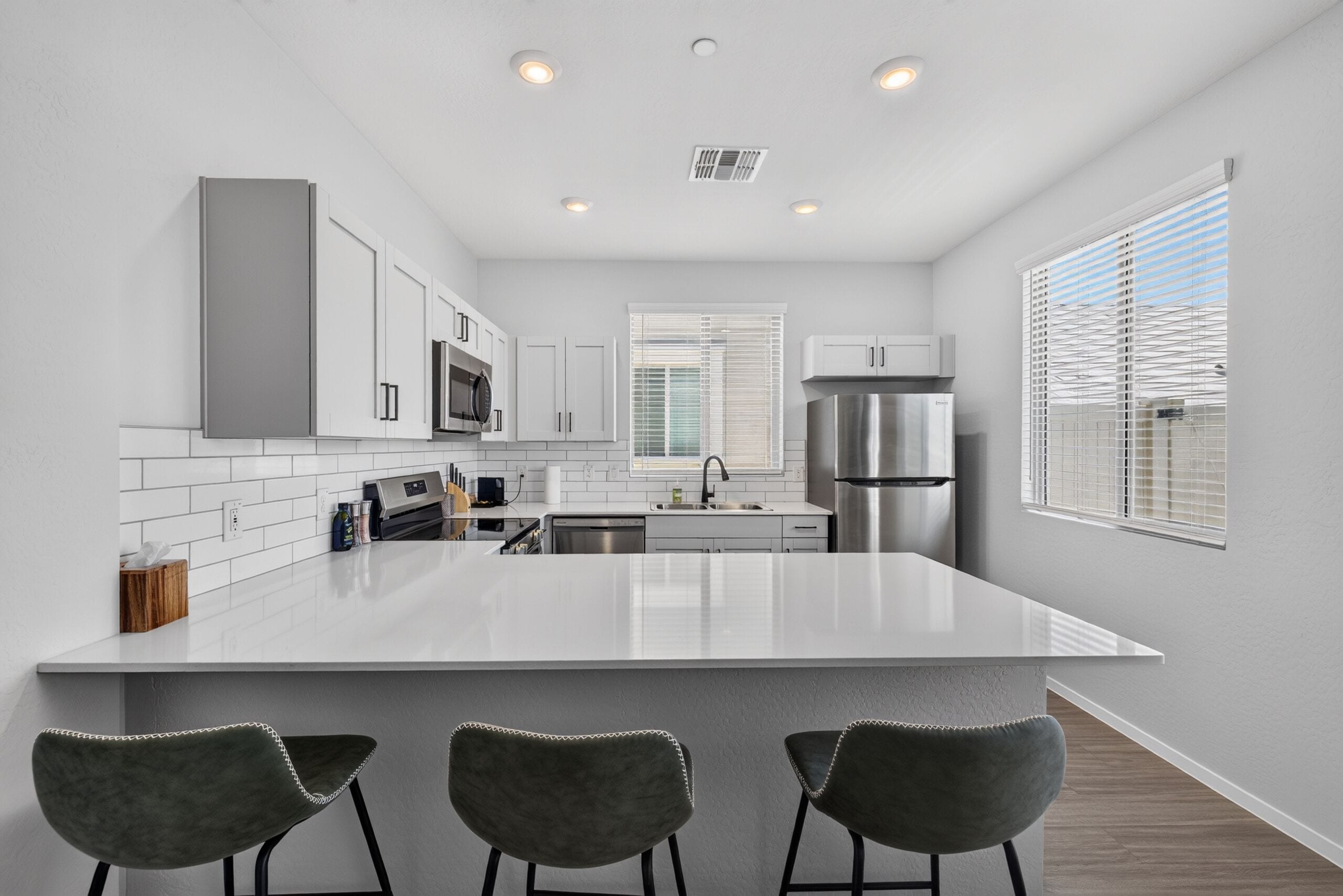 A modern kitchen with white cabinets, a stainless steel refrigerator, a large island with bar stools, and a window with blinds.