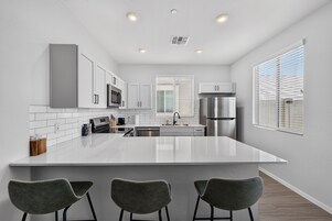 Una cocina moderna con armarios blancos, un refrigerador de acero inoxidable, una isla grande con taburetes de bar y una ventana con persianas.