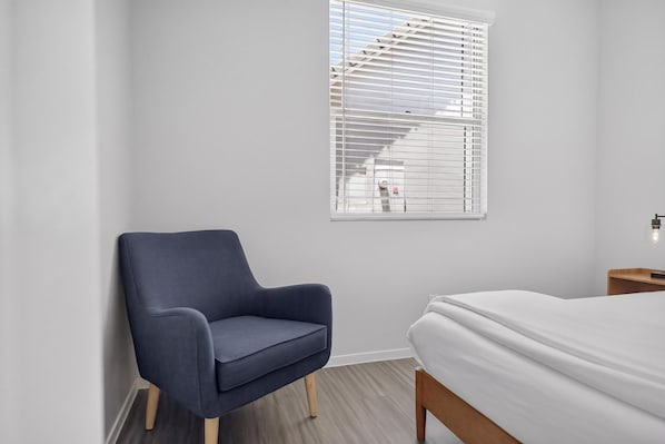 A bedroom with a bed, a chair, a window with blinds, and a nightstand.