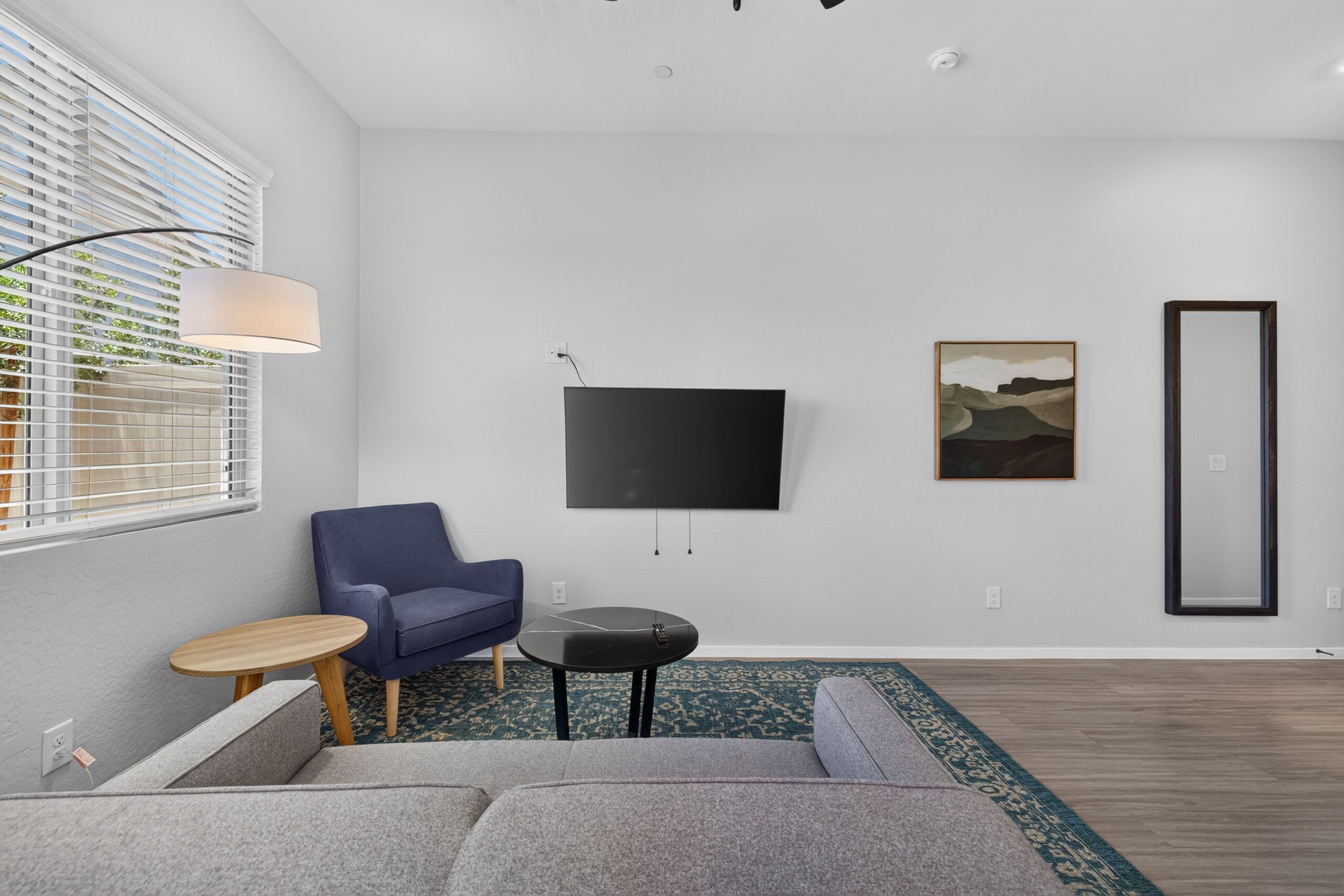 A modern living room with a grey sofa, a blue armchair, a small round table, a floor lamp, a flat-screen TV, and a framed picture on the wall.