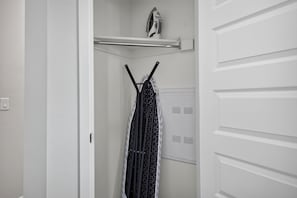 A white closet with a hanging rack, a shelf with an iron, and a door with a handle.