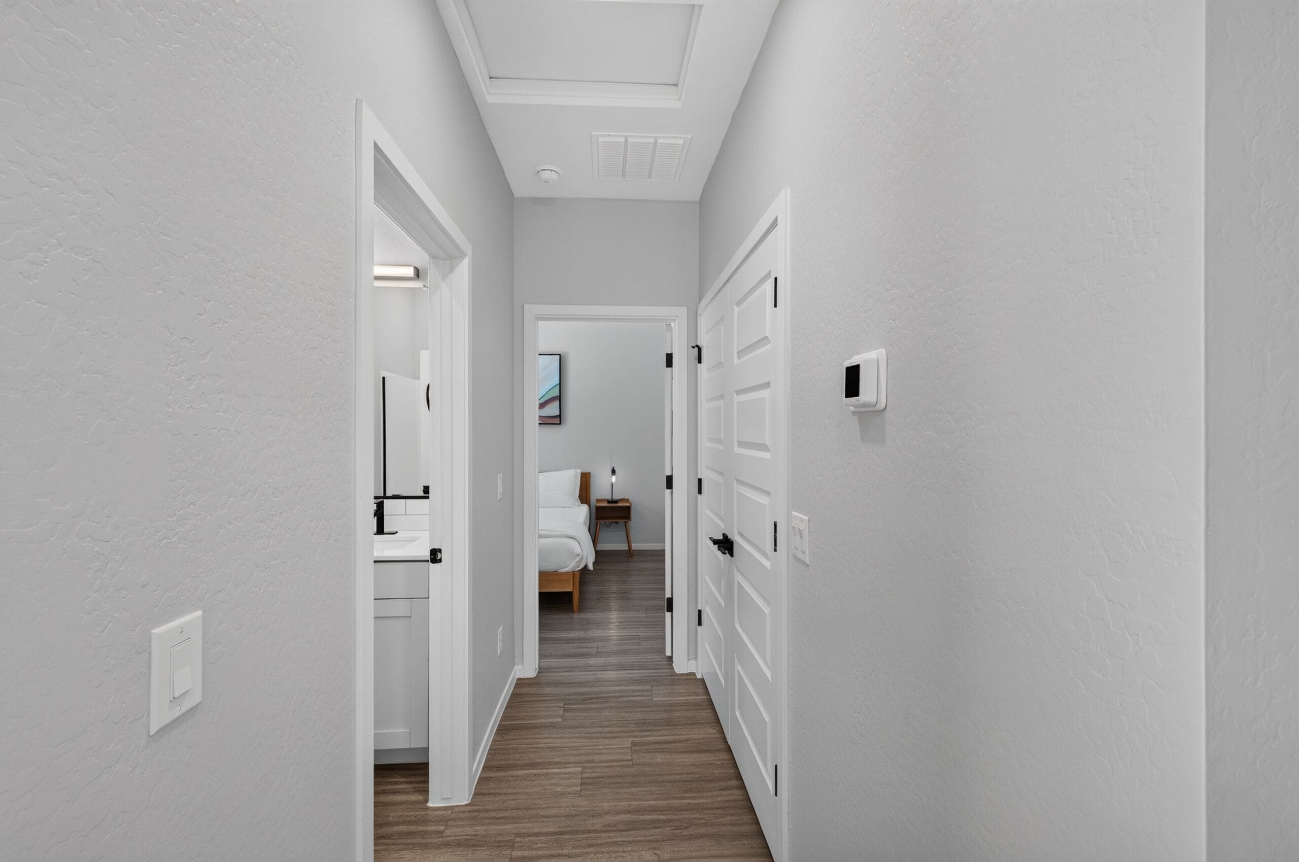 Un couloir aux murs blancs, doté d’un plancher en bois et de portes blanches donnant sur une chambre à coucher.