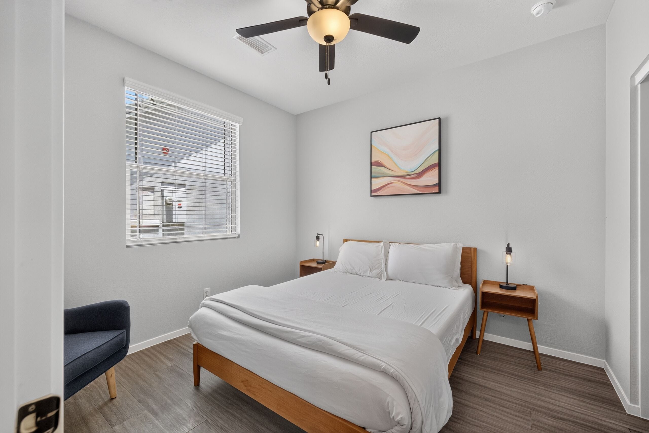 Quarto com cama, ventilador de teto, janela com persianas, mesa de cabeceira com abajur e um quadro abstrato pendurado na parede.