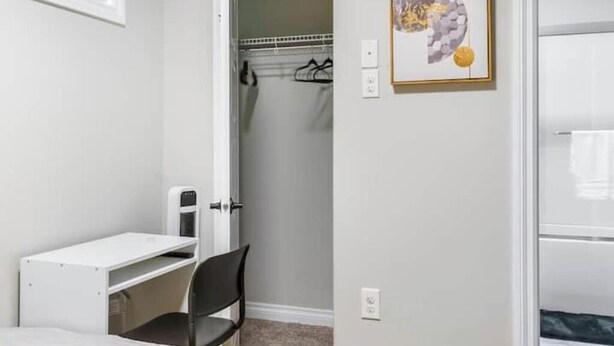 Una habitación con un escritorio blanco, una silla, una puerta que da a una ducha y un cuadro abstracto enmarcado en la pared.