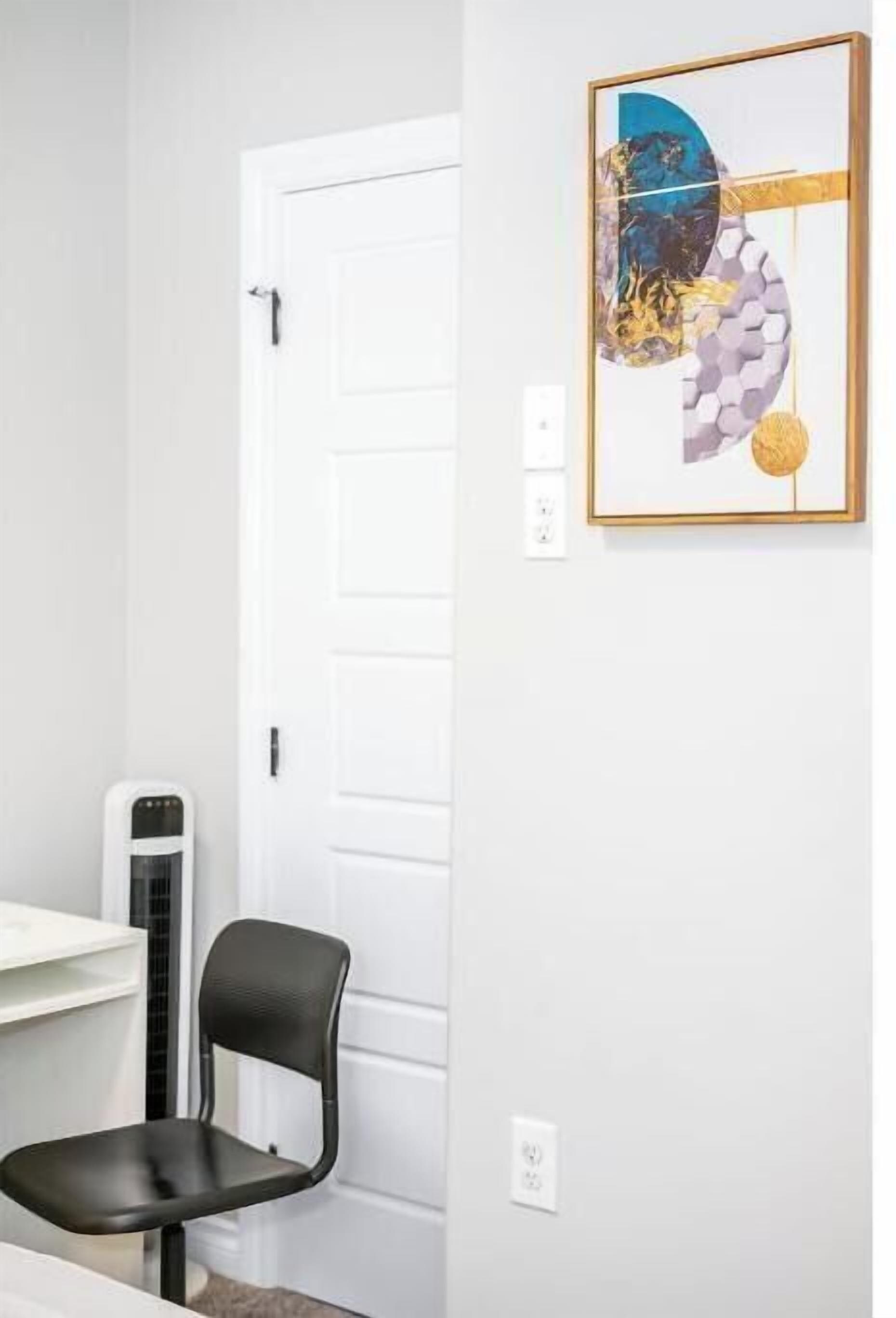 Une pièce avec une porte blanche, une chaise noire, un bureau blanc et un tableau abstrait encadré accroché au mur.