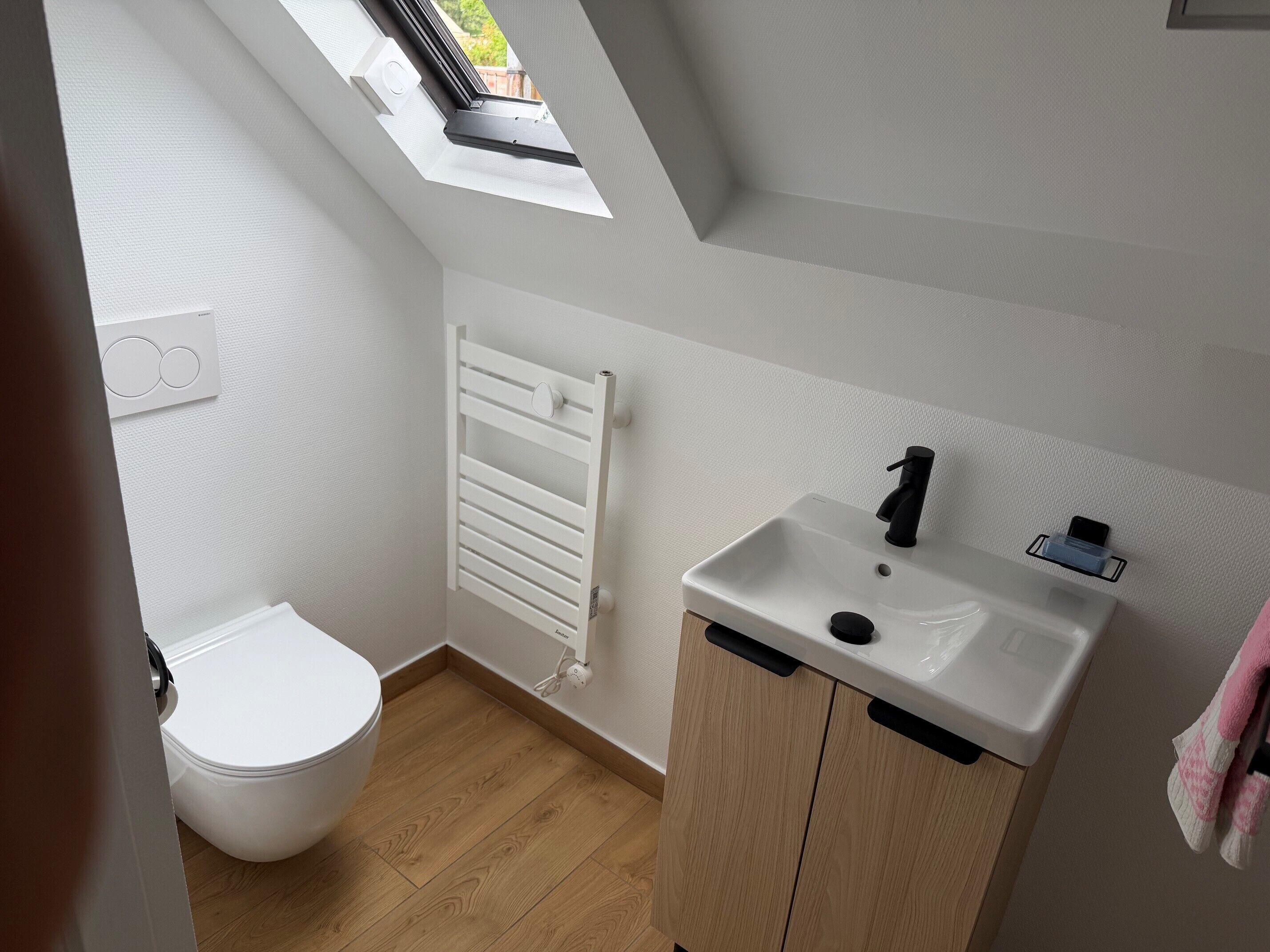 Ein kleines, modernes Badezimmer mit Dachfenster, einem weißen Heizkörper, einem Waschbecken mit schwarzem Wasserhahn und einer Toilette.