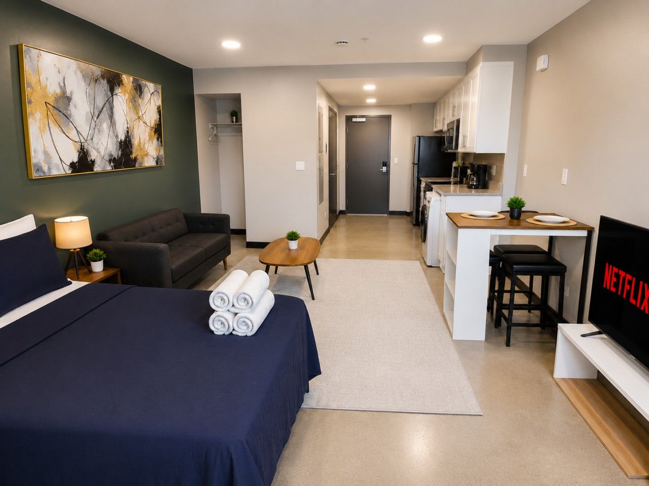 A hotel room with a bed, a desk, a kitchenette, a TV, and a painting on the wall.