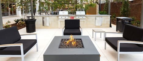 An outdoor patio with a central fire pit, black cushioned seating, and a barbecue grill.