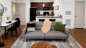 A modern living room with a grey sofa, black chairs, a dining table, and a kitchen in the background. - Iron City Retreat | Modern 2BR Stay #4 (Birmingham)