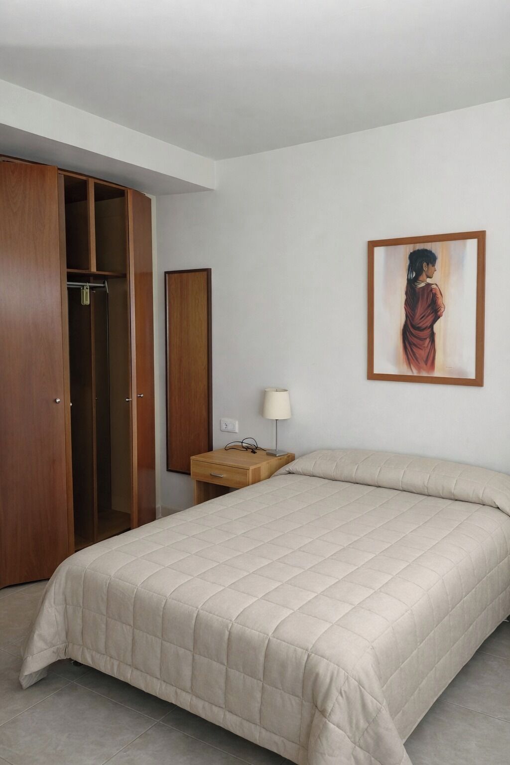 A bedroom with a bed, a wardrobe, a nightstand, a lamp, and a framed picture on the wall.