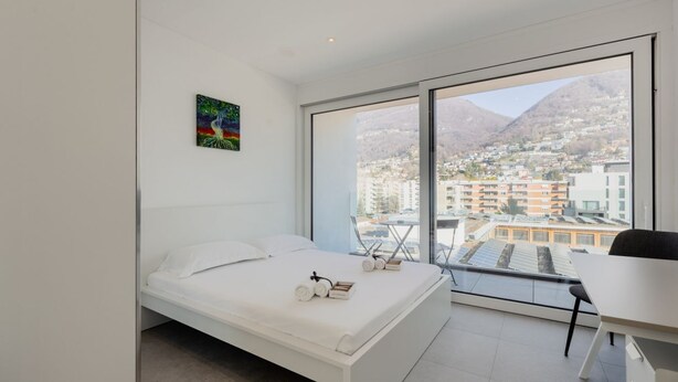 A modern bedroom with a bed, a desk, and a large window offering a city view. - {Suite 507} Gym, Business Room, Modern, Relax (Lugano)