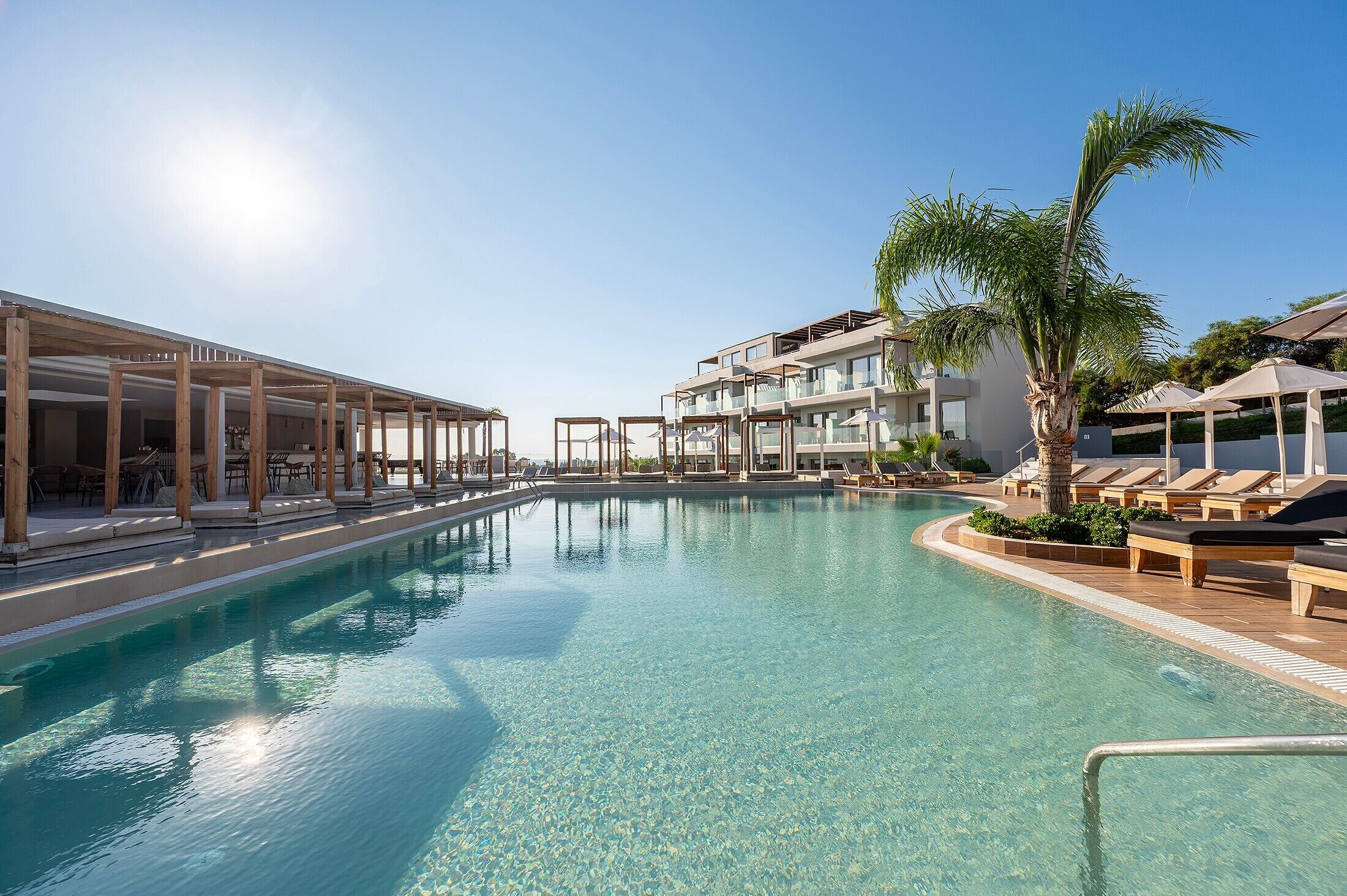 Una piscina cristalina con sillas de descanso y sombrillas, rodeada de edificios modernos y palmeras.