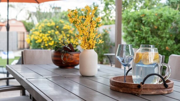 Une table en bois sur laquelle se trouve un vase de fleurs jaunes, une carafe en verre avec une tranche de citron et un verre à vin.