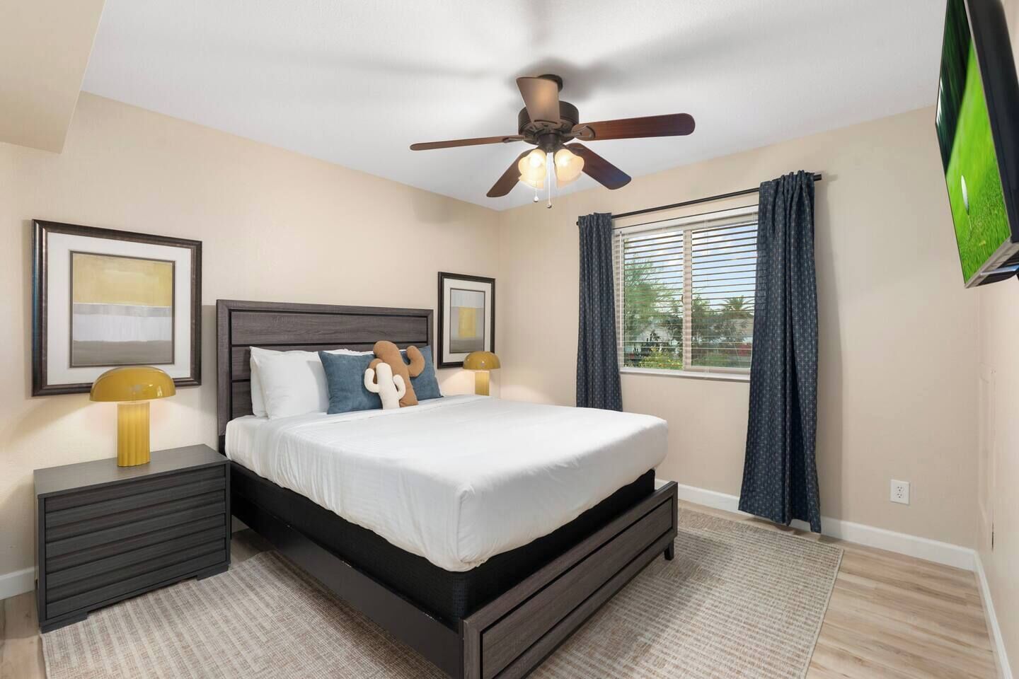 A bedroom with a bed, a ceiling fan, a nightstand, a lamp, a window with curtains, and a TV mounted on the wall.