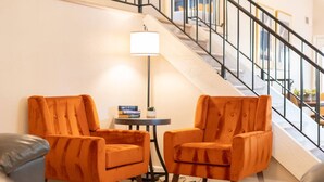Un intérieur moderne avec deux fauteuils orange, une petite table ronde et un lampadaire. Un escalier avec une rampe en métal est visible au fond.