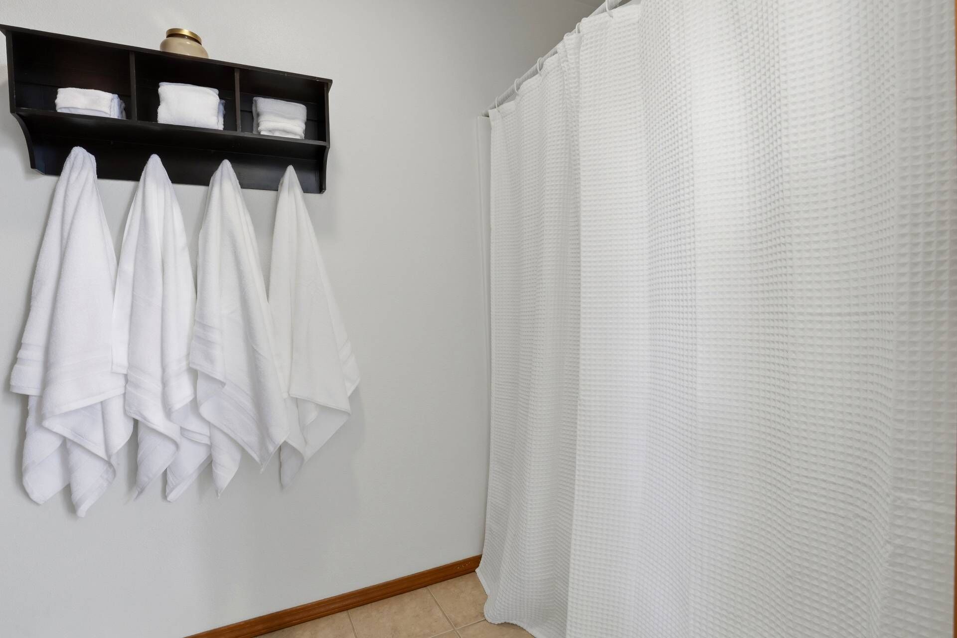 A bathroom with a shower curtain, a towel rack with several towels, and a shelf with a vase.