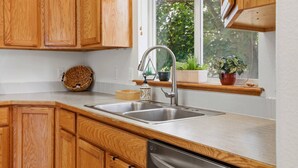 A kitchen with wooden cabinets, a stainless steel sink, a built-in dishwasher, and a window with plants on the windowsill.