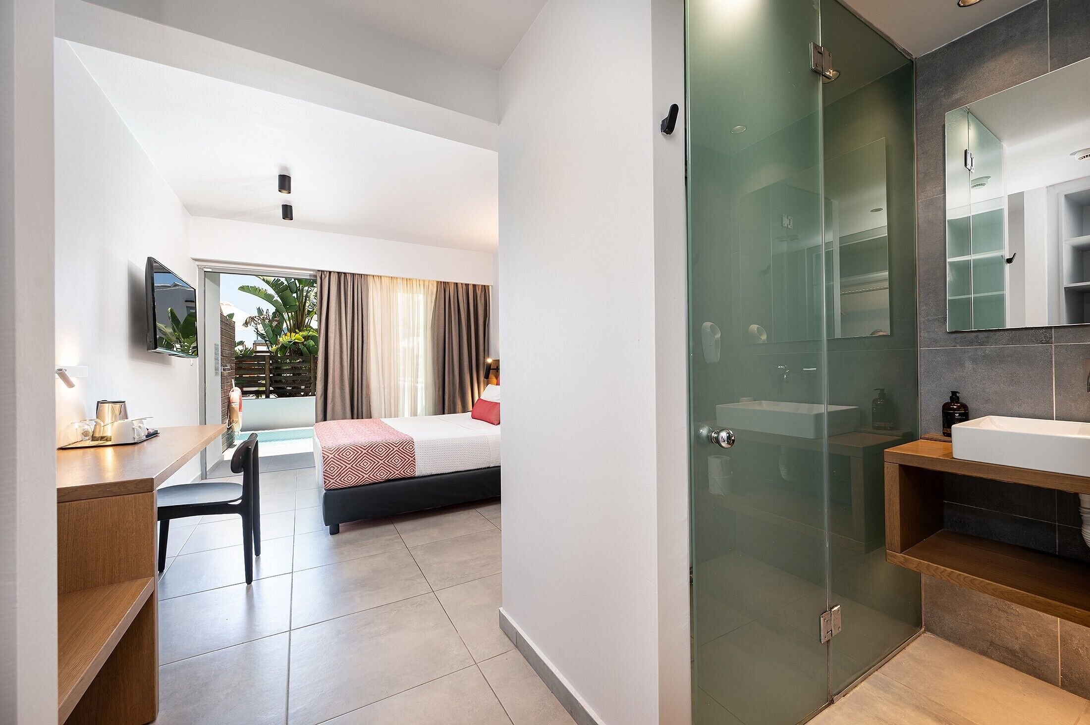 Ein Hotelzimmer mit Bett, Schreibtisch, Stuhl und einem Badezimmer mit Duschkabine aus Glas. An der Wand ist ein Fernseher angebracht.