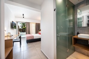 Ein Hotelzimmer mit Bett, Schreibtisch, Stuhl und einem Badezimmer mit Duschkabine aus Glas. An der Wand ist ein Fernseher angebracht.