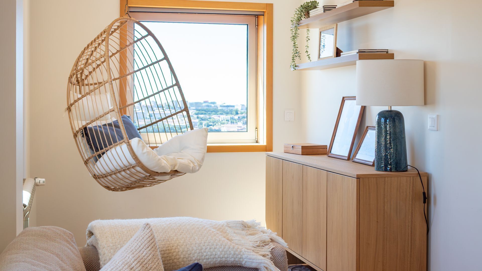 A room with a hanging chair, a window with a view, a cabinet with a lamp, and a bed with a blanket.