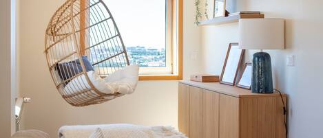 A room with a hanging chair, a window with a view, a cabinet with a lamp, and a bed with a blanket.