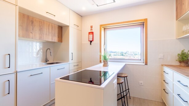Una cocina moderna con muebles blancos, una placa de inducción negra, un ventanal con vistas y un extintor rojo en la pared.