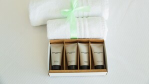 A neatly folded white towel with a light green ribbon tied around it, placed on a bed next to a box containing four toiletry items. - {Suite 510} Gym, Business Room, Modern, Relax (Lugano)