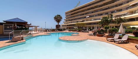 A hotel with a large swimming pool, sun loungers, and a palm tree.