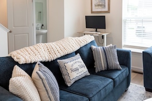 A living room with a blue sofa, a cream-colored throw blanket, and striped pillows. A framed quote is visible on one pillow. - 13 Min to Beaver Stadium | Game Room, Deck (Bellefonte)