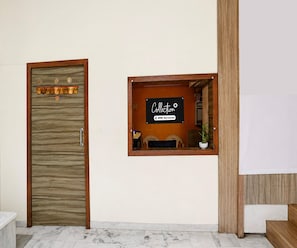 Un área de recepción con una puerta de madera, un cartel que indica 'Collection' y 'OYO-Serviced', y una ventana pequeña con un escritorio y una silla.
