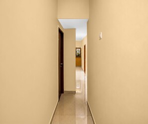 A corridor with beige walls and a tiled floor, leading to a room with a door and a window with a flower decoration. - Classic Hotel Lal Sai Residency (Mumbai)