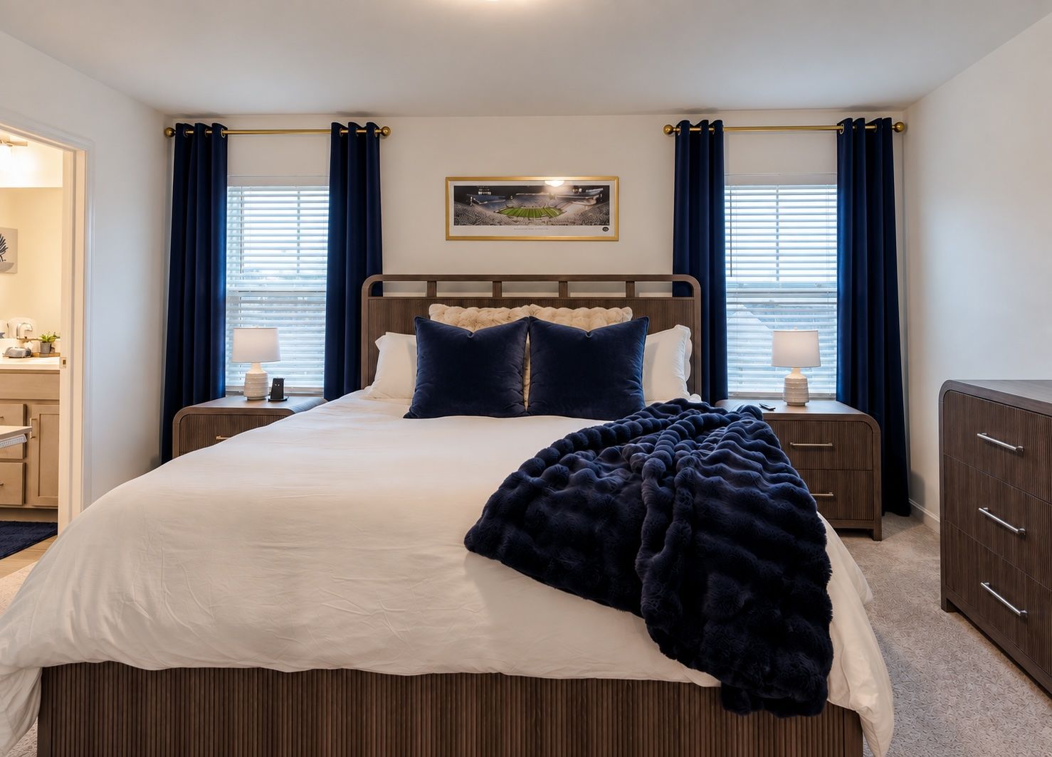 A bedroom with a large bed, two bedside tables, a headboard, a framed picture on the wall, and a window with curtains.