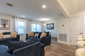 A modern living room with a dark blue sofa set, a large window with white curtains, a pendant light, and a framed picture on the wall.