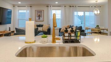 A modern kitchen with a stainless steel sink, a gold faucet, and a white countertop. There is a dining area with a table and chairs, and a living room with a sofa and a TV.