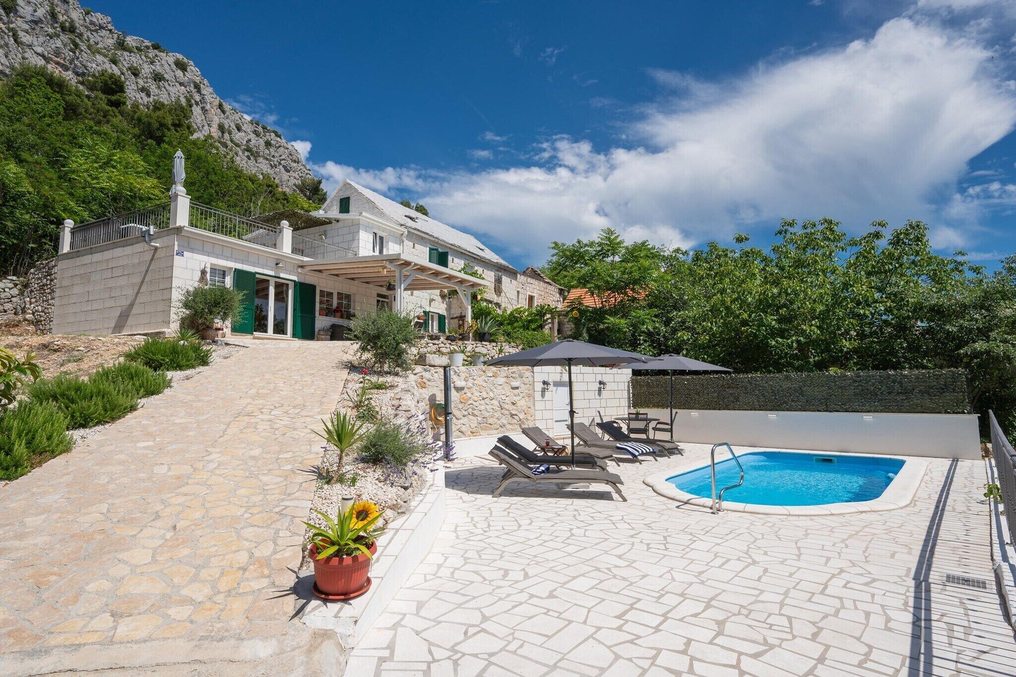 A stone-paved outdoor area with a swimming pool, lounge chairs, and a house with a balcony.