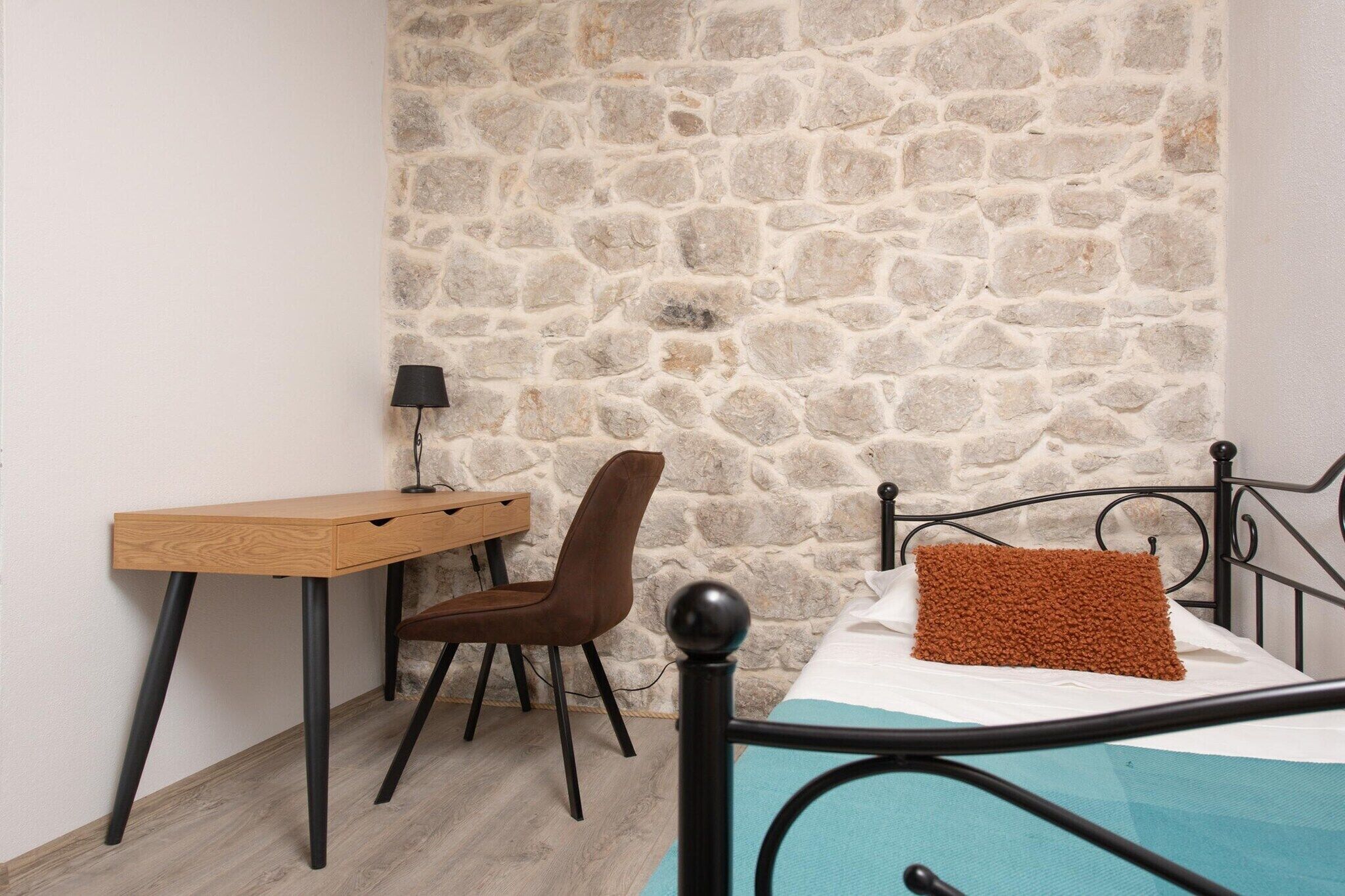 Ein Schlafzimmer mit einer Steinwand, einem Holztisch, einem braunen Stuhl, einem Bett mit blauer Matratze und einem schwarzen Metall-Kopfende.